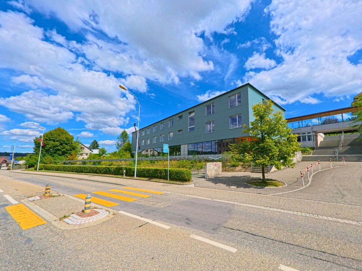 Ein modernes Schulgebäude mit Treppenanlage und Fussgängerstreifen liegt an einer leicht ansteigenden Strasse in Safenwil (5745, Kanton Aargau) bei sonnigem Wetter.