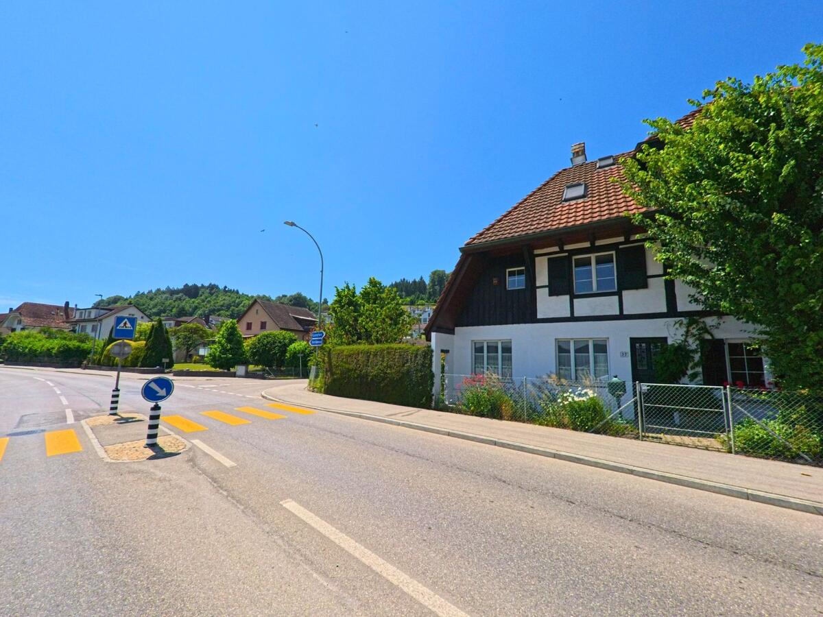 Am Rand einer Kurve mit Verkehrsinsel in Safnern (2553, Kanton Bern) steht ein traditionelles Wohnhaus mit dunklem Fachwerk und gepflegtem Garten.