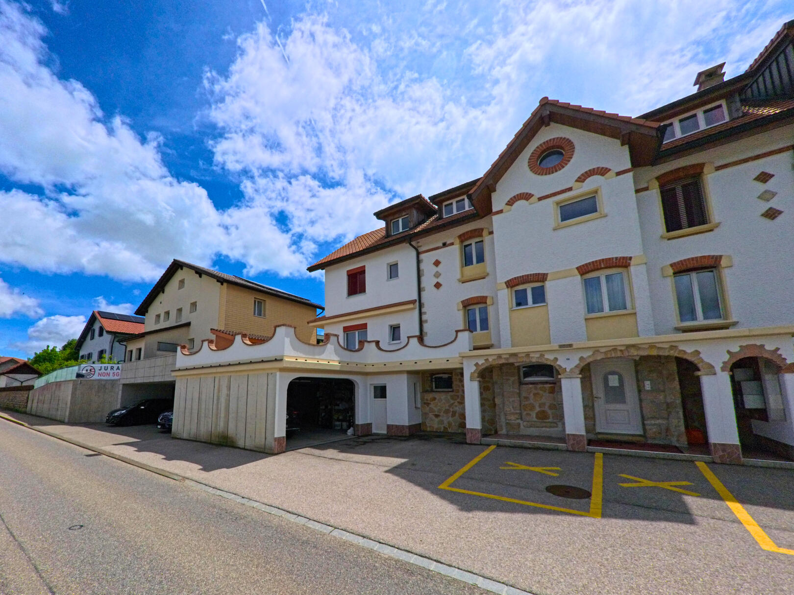 In Saignelégier (2350, Kanton Jura) ist eine Strasse zu sehen, die an einem Gebäude mit auffälliger Architektur und Parkplätzen vorbeiführt.