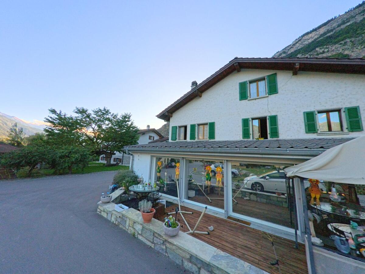 Ein traditionelles Wohnhaus mit gruenen Fensterlaeden steht in Saillon (1913, Kanton Wallis) neben einer kleinen Terrasse mit Dekorationen.