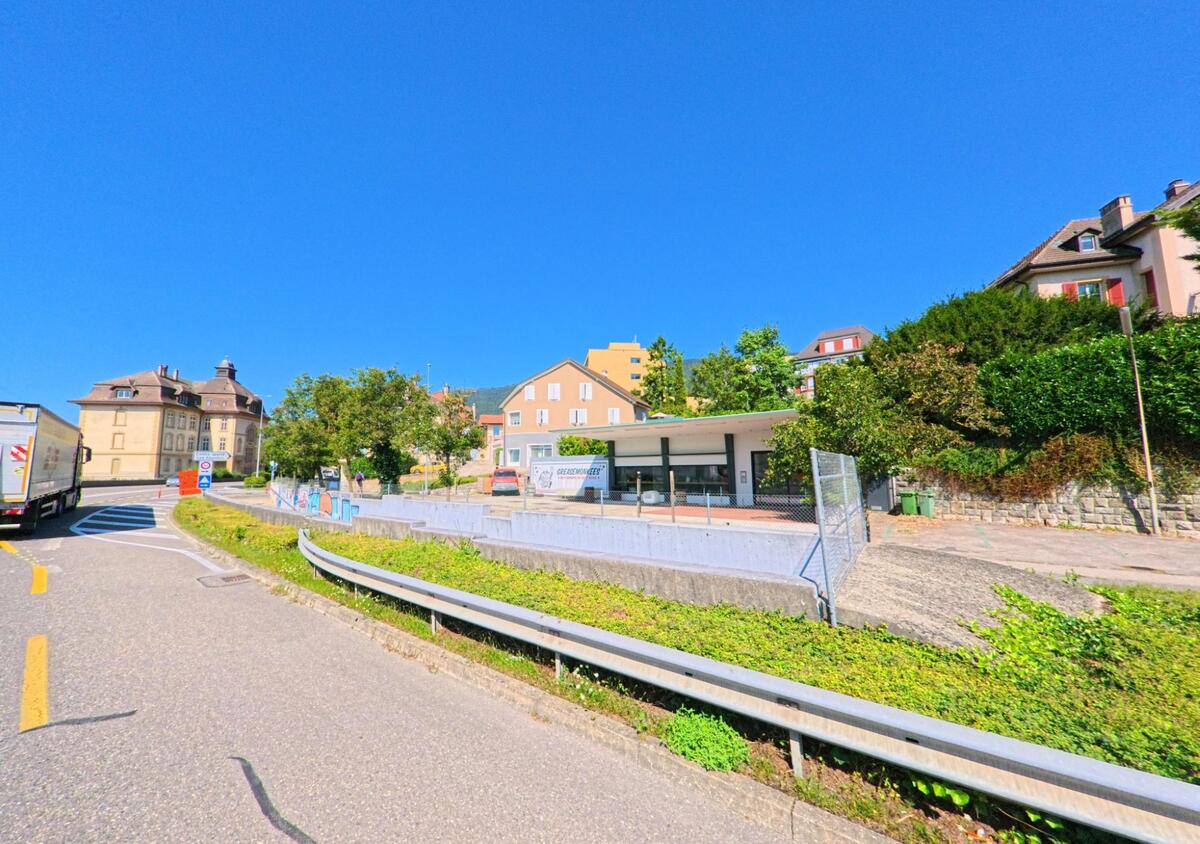 In Saint-Blaise (2072, Kanton Neuenburg) fuehrt eine Strasse an einem kleinen modernen Pavillon vorbei, waehrend im Hintergrund historische Gebaeude und Wohnhaeuser stehen.