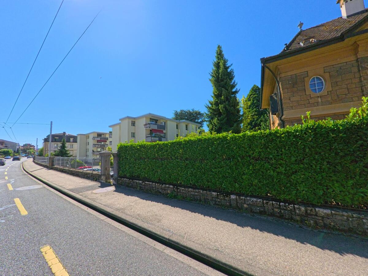 Eine von Wohnhaeusern gesaeumte Strasse in Saint-Blaise (2072, Kanton Neuenburg) verlaeuft unter strahlendem Sonnenschein, teils von einer hohen Hecke begrenzt.
