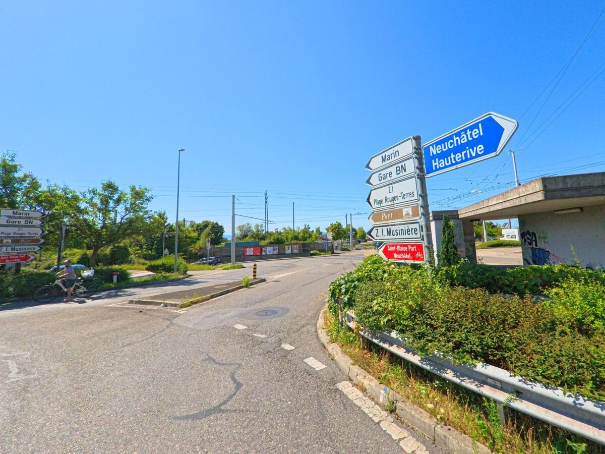 An einer Strassenabzweigung in Saint-Blaise (2072, Kanton Neuenburg) weisen mehrere Wegweiser zu umliegenden Orten und zum Hafen, waehrend im Hintergrund Bahngleise verlaufen.