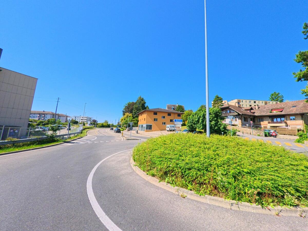 Eine begruente Verkehrsinsel liegt in Saint-Blaise (2072, Kanton Neuenburg) an einer leicht ansteigenden Strasse mit Wohn- und Geschaeftsgebaeuden im Hintergrund.