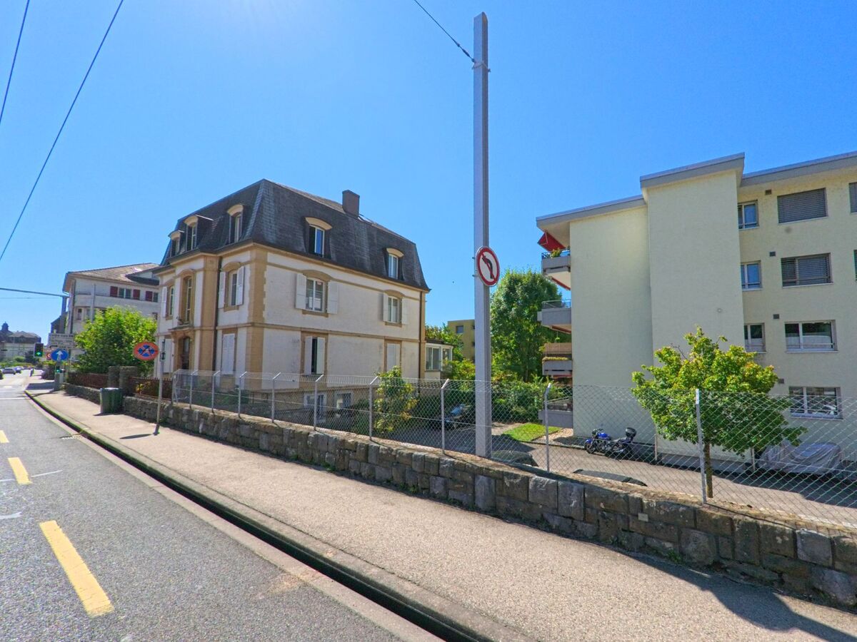 In Saint-Blaise (2072, Kanton Neuenburg) steht eine historische Villa mit Mansarddach neben einem modernen Mehrfamilienhaus an einer leicht ansteigenden Strasse.