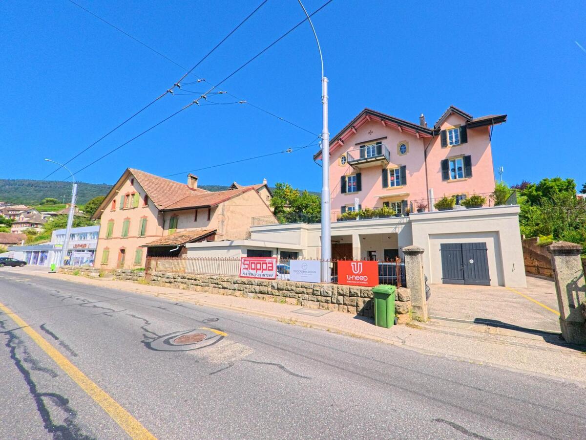 Zwei gepflegte Wohnhaeuser mit unterschiedlichen Fassadenstilen stehen in Saint-Blaise (2072, Kanton Neuenburg) an einer Strasse mit Blick auf die Rebhuegel.