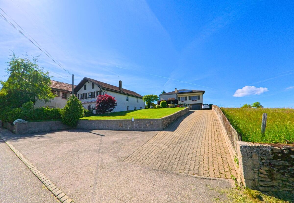 In Saint-Brais (2364, Kanton Jura) führt eine gepflasterte Straße zu einem modernen Gebäude, umgeben von grünem Gras und Bäumen.