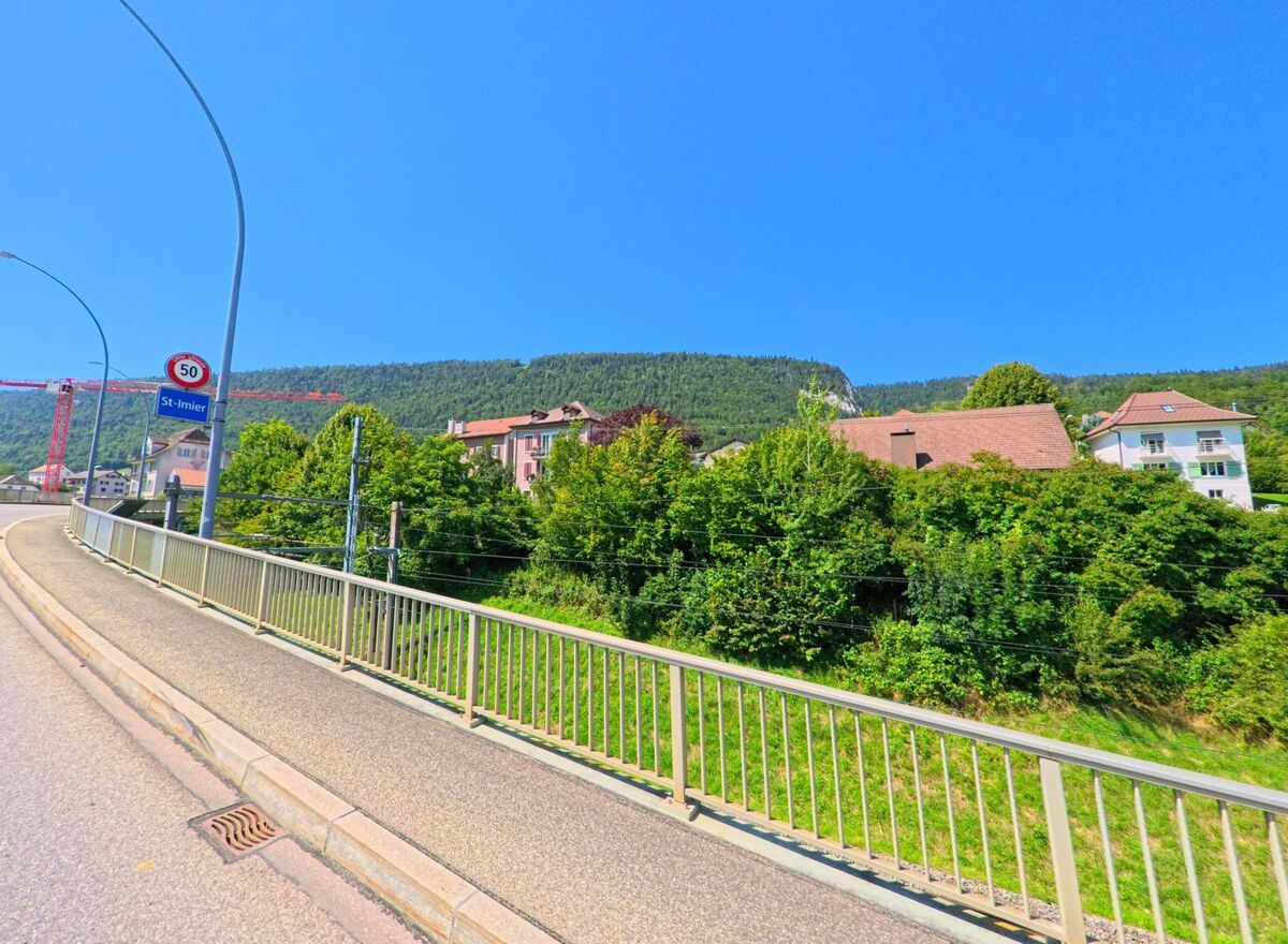 Am Ortseingang von Saint-Imier (2610, Kanton Bern) fuehrt eine Strasse mit Gehweg und Geländer an Wohnhaeusern und gruener Hanglandschaft vorbei.