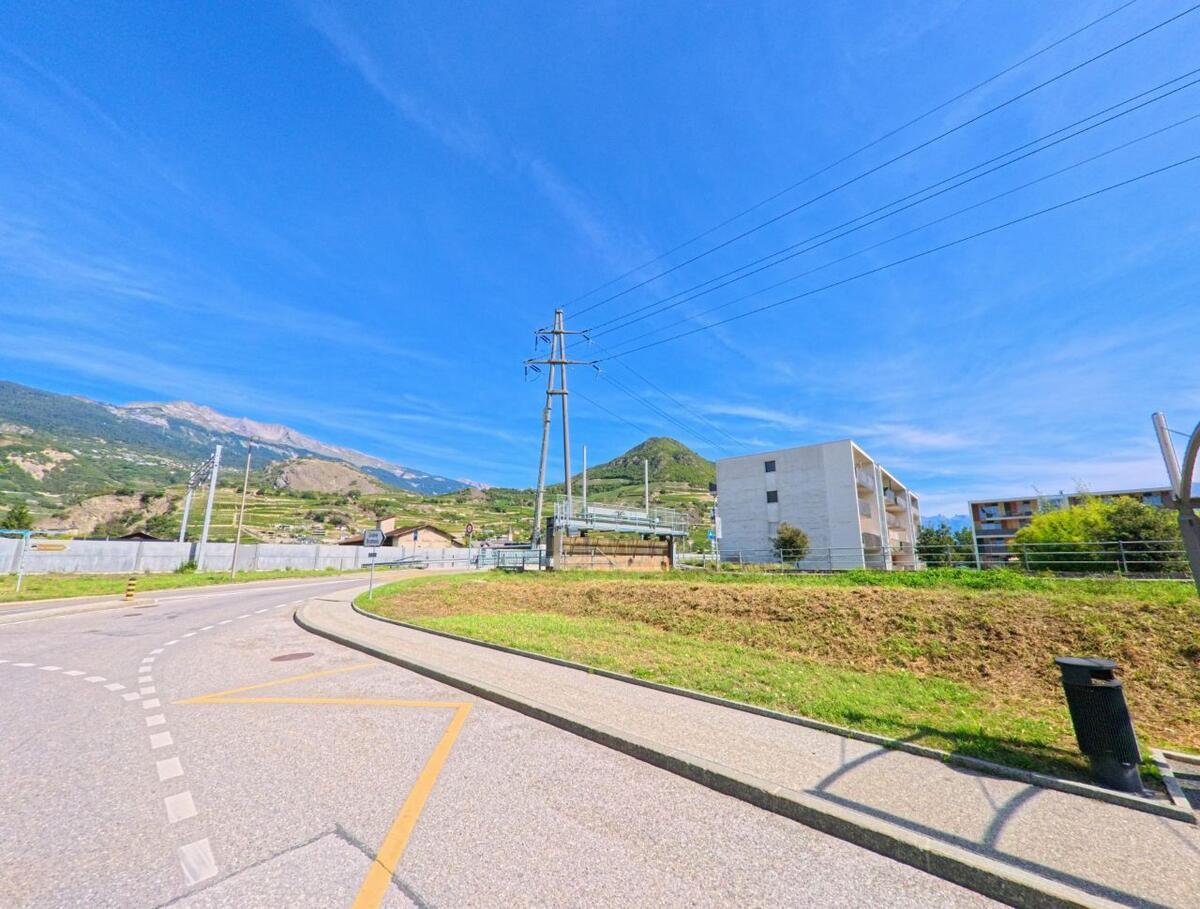 Eine kurvige Strasse in Saint-Léonard (1958, Kanton Wallis) fuehrt an modernen Wohnbauten vorbei, waehrend Stromleitungen ueber das Tal verlaufen.
