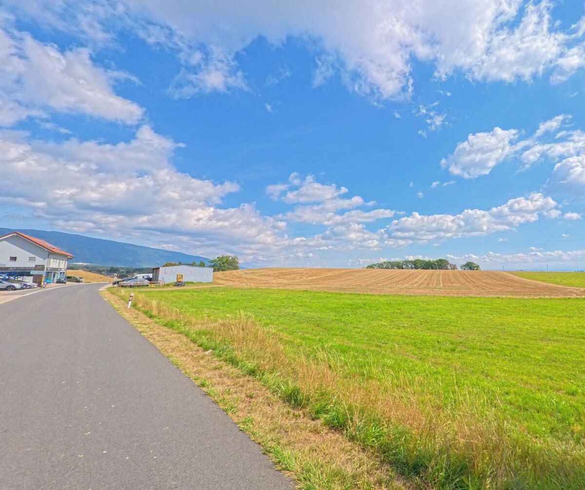 Eine Strasse führt an weiten Feldern vorbei, während am Rand von Saint-Oyens (1187, Kanton Waadt) ein modernes Gebäude und ein Lagerplatz stehen.
