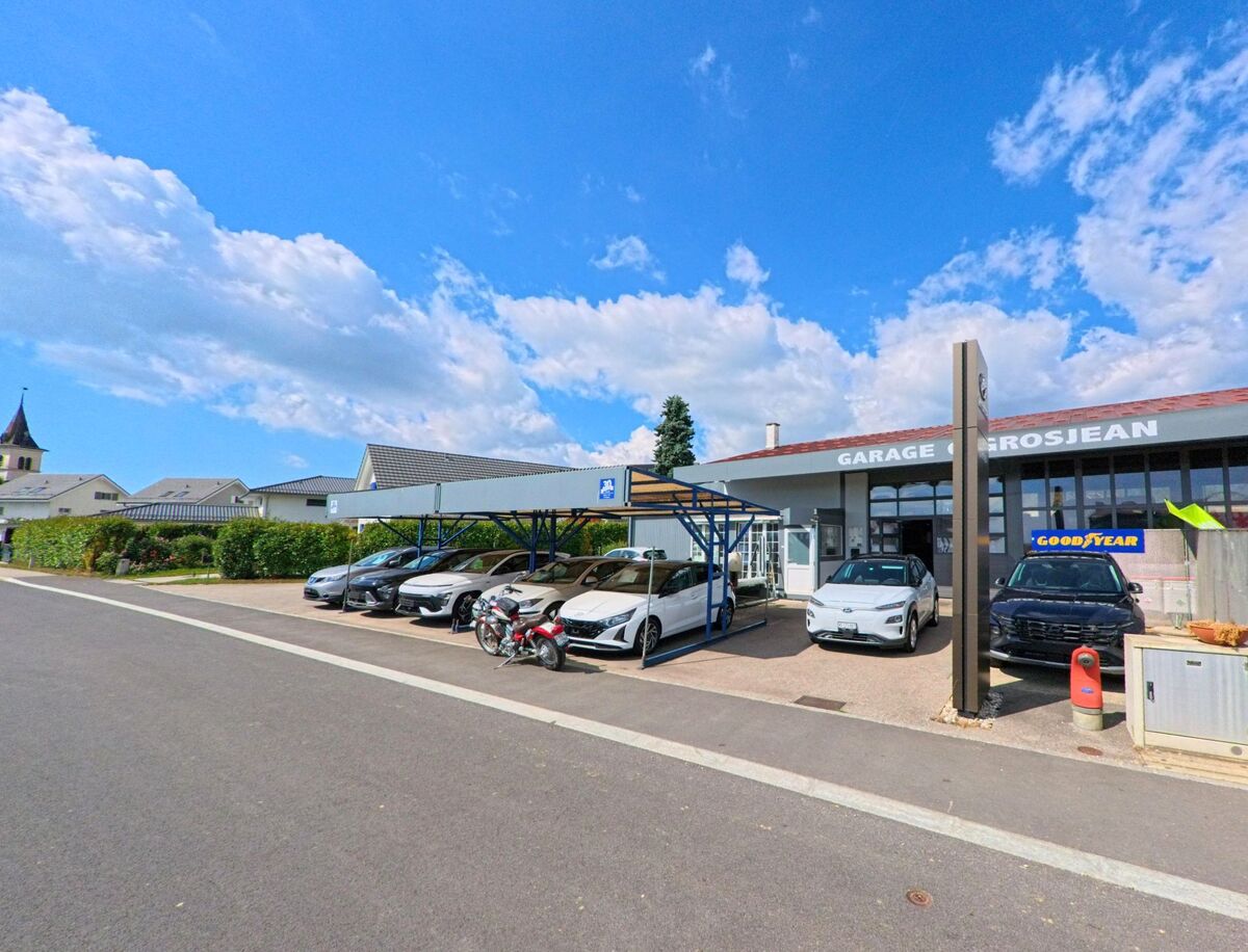 Vor der Garage Grosjean in Saint-Oyens (1187, Kanton Waadt) stehen mehrere Autos und Motorräder, während die Kirche im Hintergrund sichtbar ist.