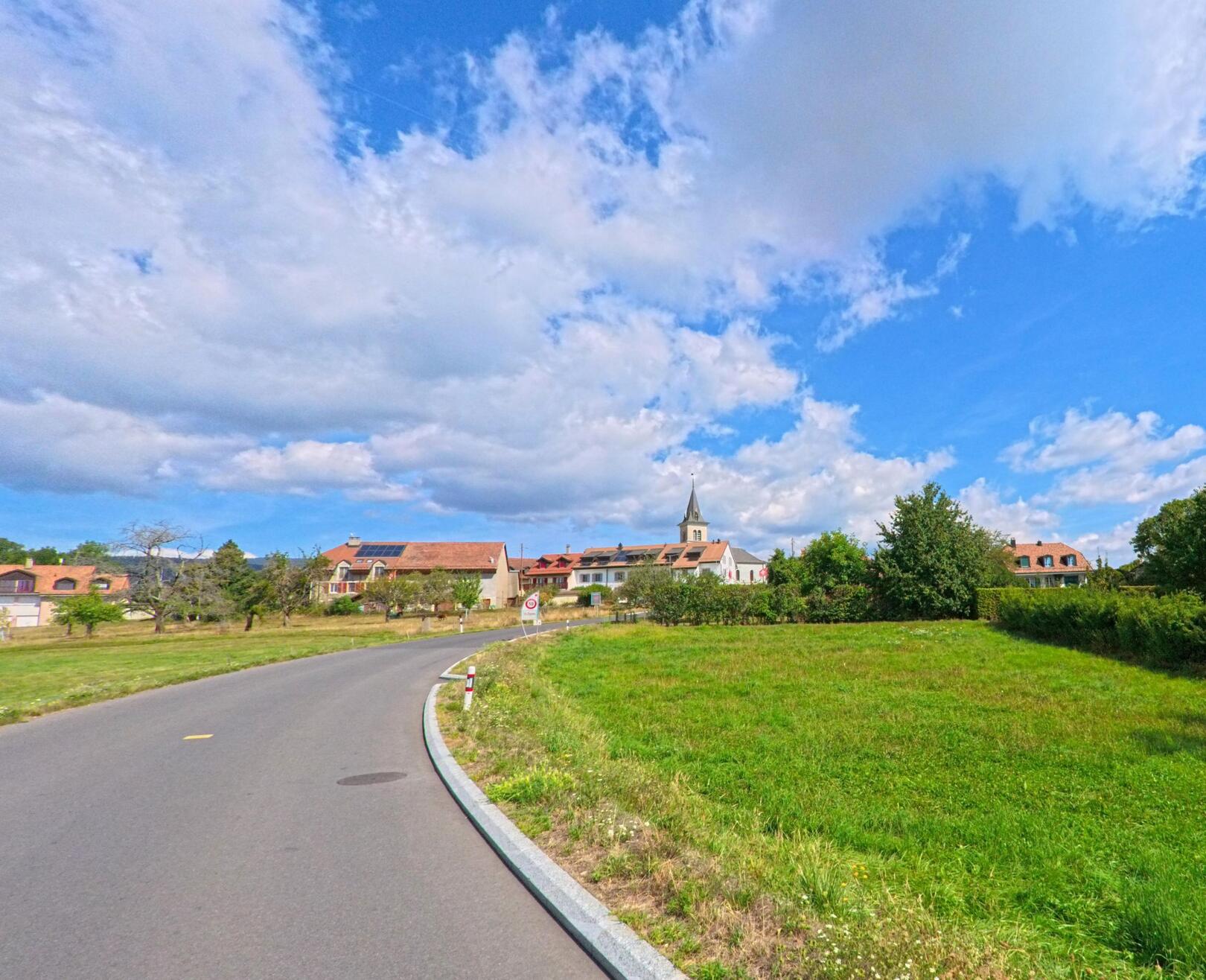 Eine Landstrasse führt durch grüne Felder auf das Dorf Saint-Oyens (1187, Kanton Waadt) mit sichtbarem Kirchturm im Hintergrund zu.