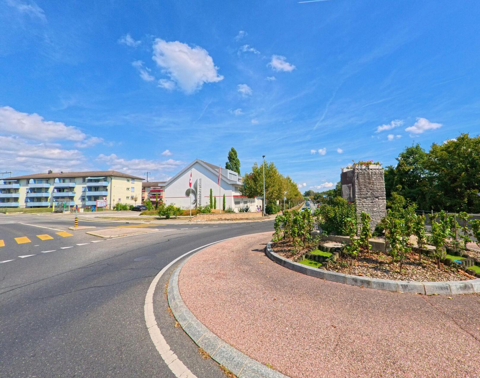 Ein bepflanzter Kreisel mit Steinmauer prägt das Strassenbild in Saint-Prex (1162, Kanton Waadt), im Hintergrund stehen moderne Wohnhäuser und ein weisses Gebäude.