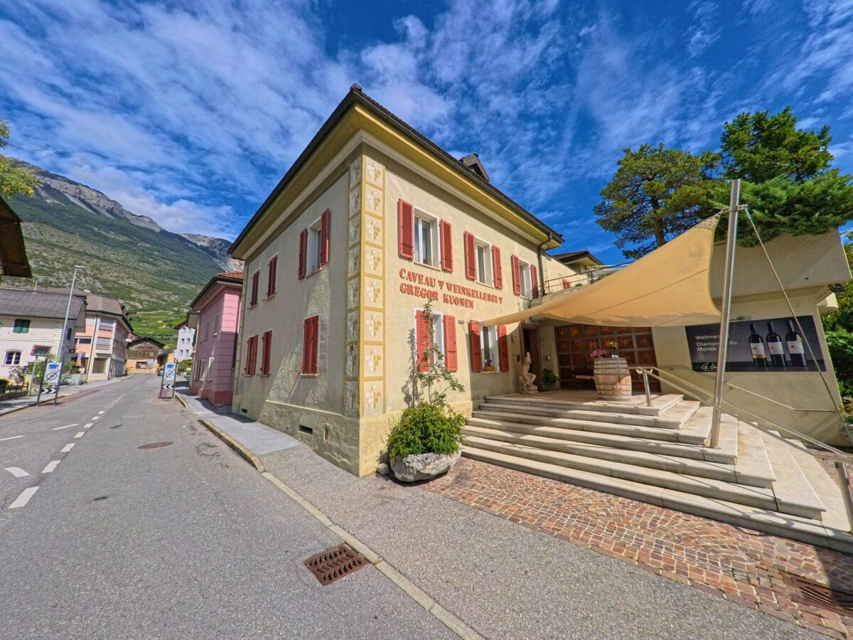 Ein traditionsreiches Gebaeude mit roten Fensterlaeden beherbergt eine Weinkellerei in Salgesch (3970, Kanton Wallis) und liegt an einer schmalen Strasse mit Blick auf die Rebhänge.