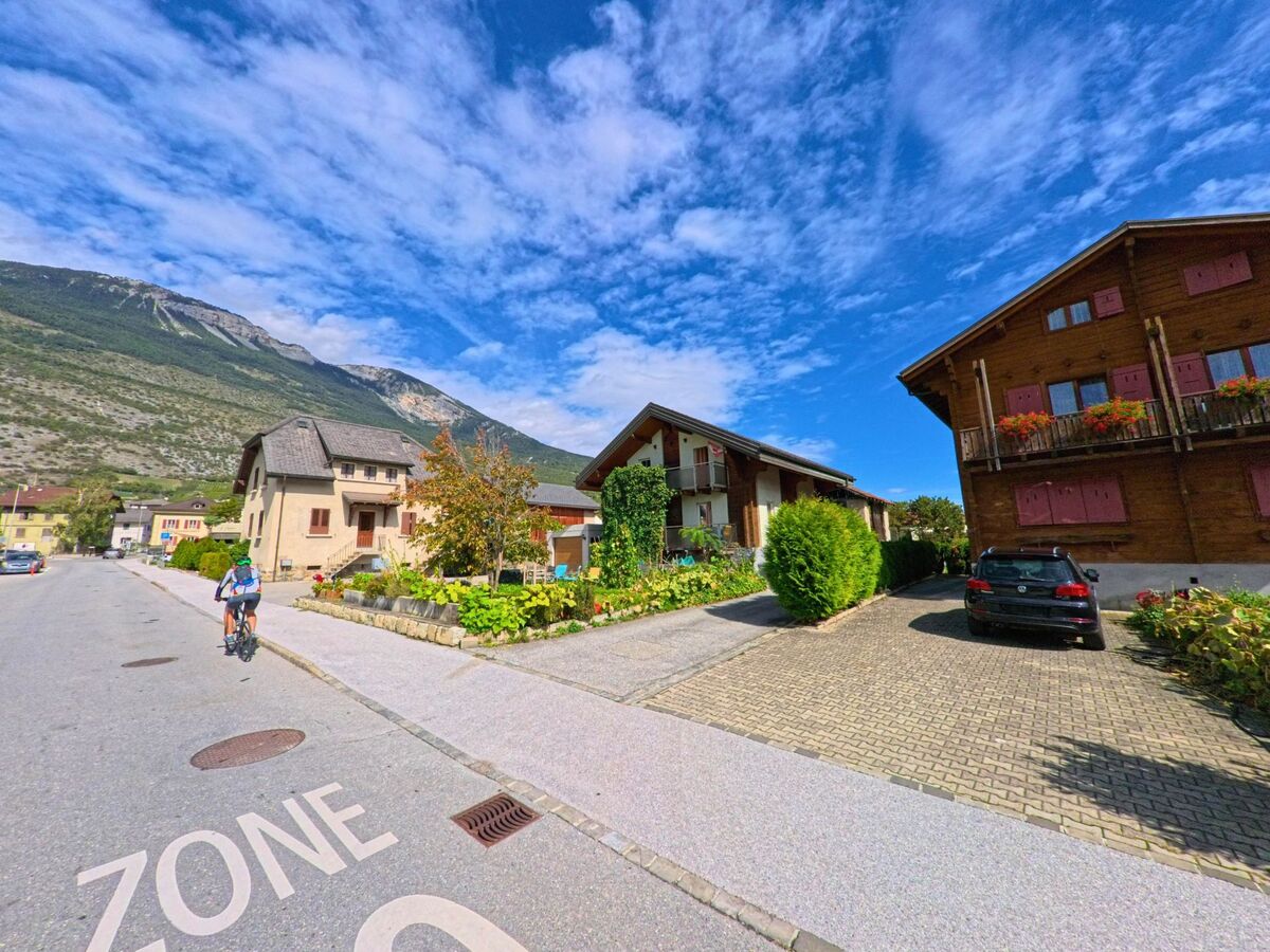 In einer Wohnstrasse von Salgesch (3970, Kanton Wallis) fahren Radfahrer vorbei an traditionellen Haeusern mit Blumenschmuck und Blick auf den nahen Berghang.