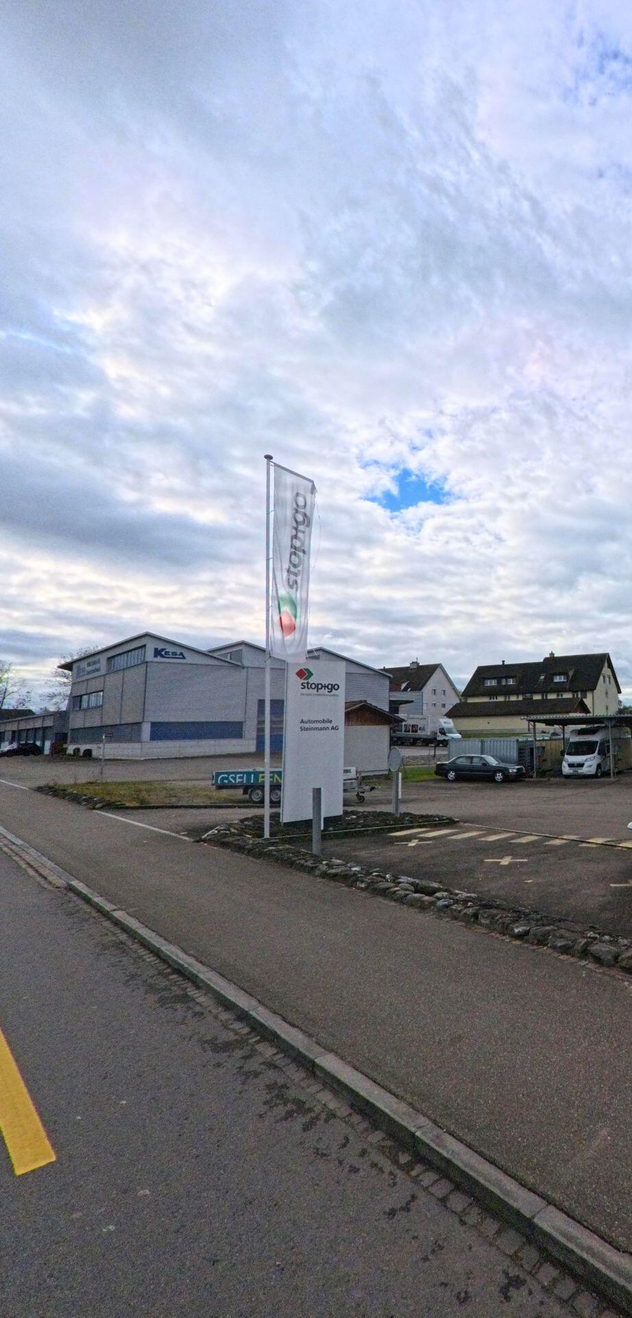 Ein Gewerbeareal mit Firmenbeschilderung, Fahne und Zufahrt liegt an einer Strasse in Salmsach (8599, Kanton Thurgau) unter aufgelockerter Bewölkung.