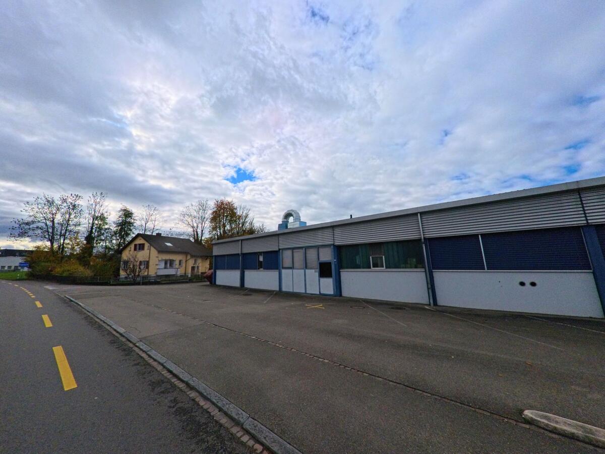 Ein langes Industriegebäude mit Vorplatz und angrenzender Fahrbahn prägt diesen ruhigen Abschnitt von Salmsach (8599, Kanton Thurgau) bei bedecktem Himmel.
