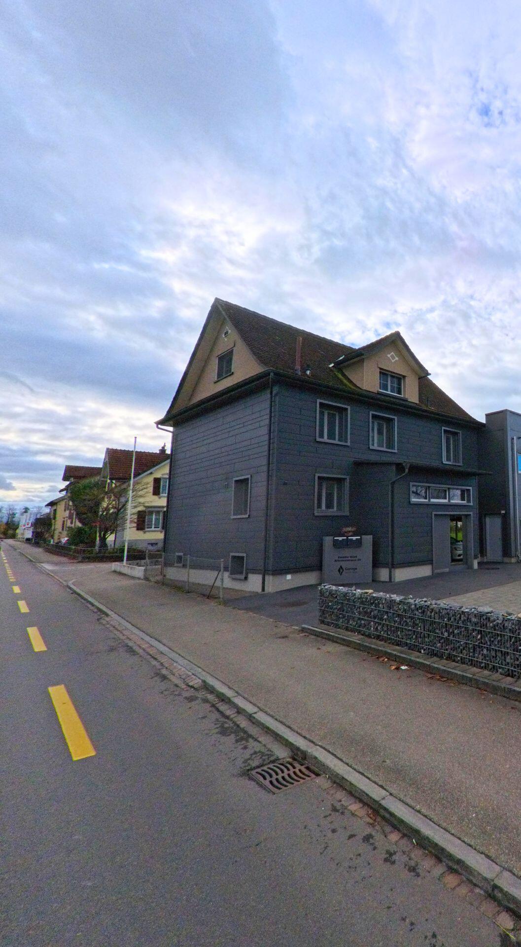 Ein graues Wohn- und Geschäftsgebäude mit Gehweg und Fahrbahn befindet sich an einer ruhigen Strasse in Salmsach (8599, Kanton Thurgau) unter leicht bewölktem Himmel.