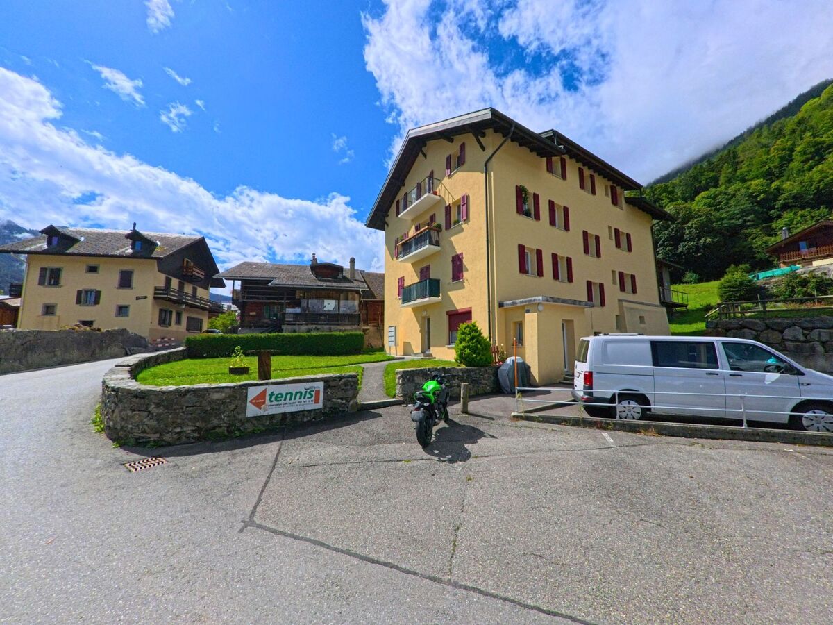 Ein gelbes Wohnhaus mit roten Fensterlaeden steht in Salvan (1923, Kanton Wallis) an einer Strasse mit Hinweisschild zum Tennisplatz.