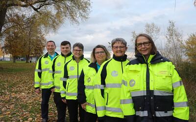 Die Mitglieder des Samaritervereins Altnau und Umgebung aus Altnau (8595, Kanton Thurgau) posieren gemeinsam in leuchtend gelben Einsatzjacken mit Reflexstreifen in einer herbstlichen Parklandschaft.