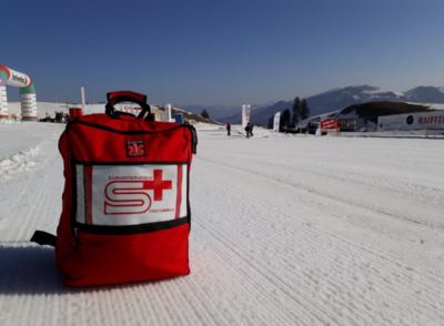 Auf einer verschneiten Flaeche steht in Zweisimmen (3770, Kanton Bern) ein roter Erste Hilfe Rucksack eines Samaritervereins vor einer winterlichen Berglandschaft.
