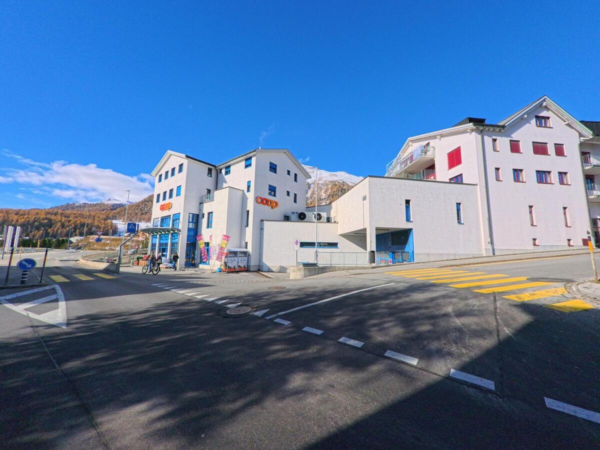 An einer Kreuzung mit Fussgängerstreifen steht ein moderner Gebäudekomplex mit Einkaufsmöglichkeit in Samedan (7503, Kanton Graubünden), umgeben von Wohnbauten und alpiner Landschaft.