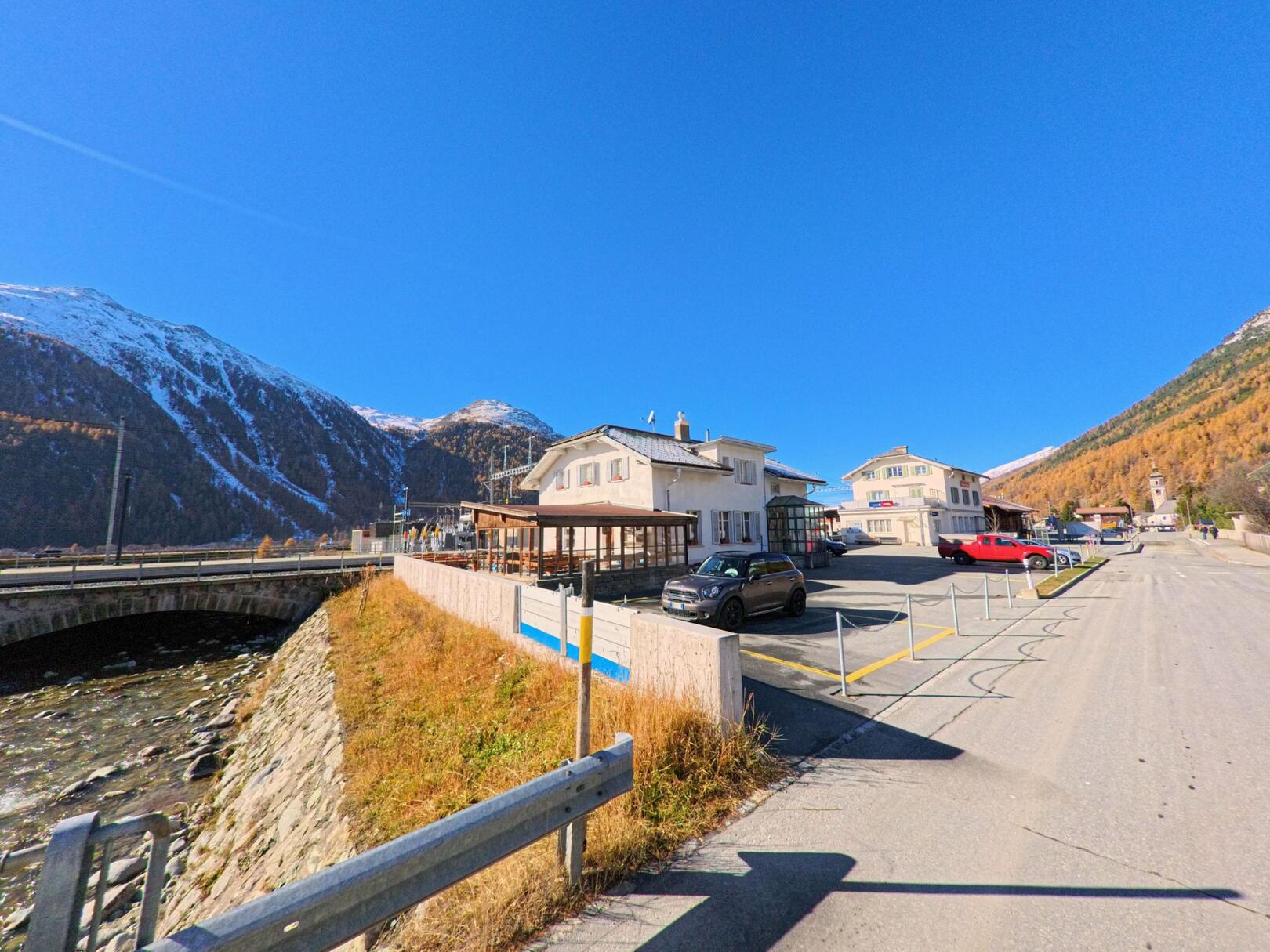 Am Rand von Samedan (7503, Kanton Graubünden) liegen Gebäude und Parkflächen nahe einer Brücke über den Fluss, eingerahmt von alpinen Hängen und offenem Himmel.