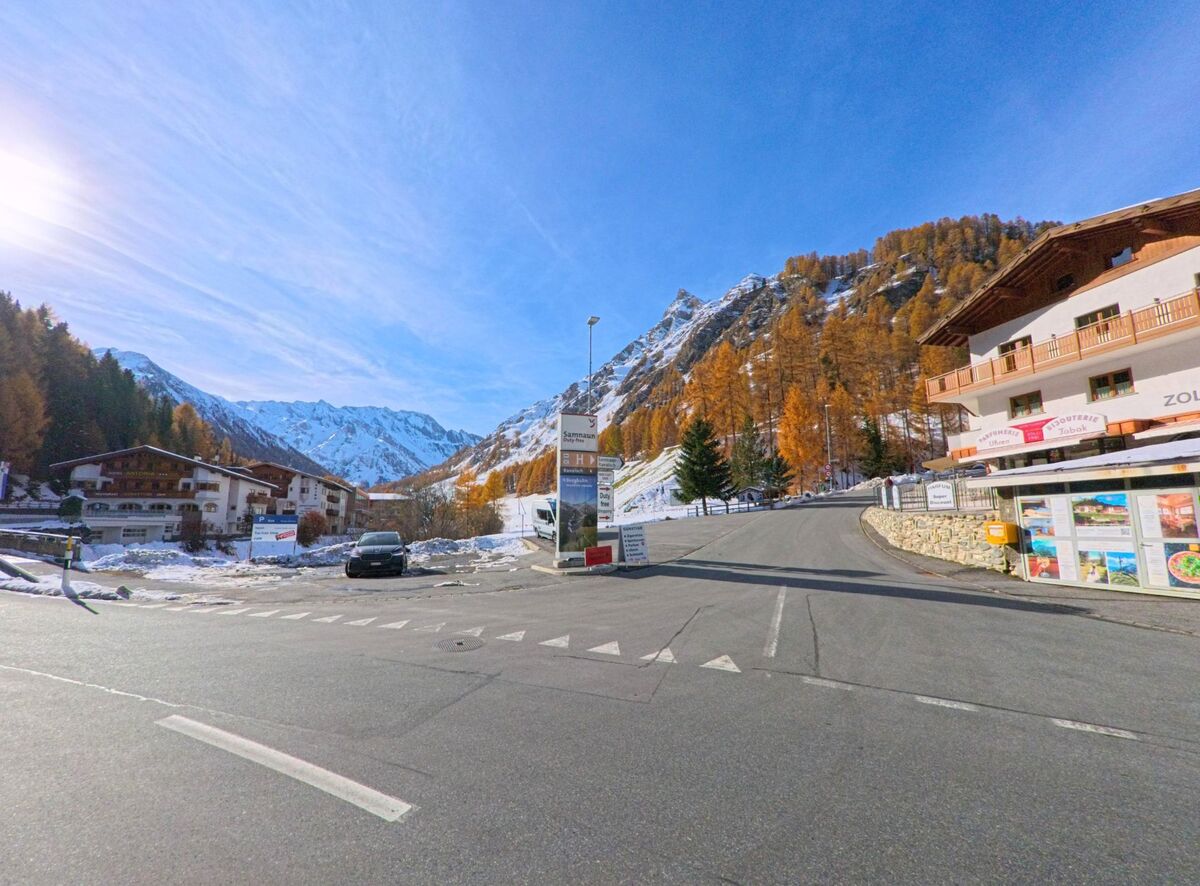 An einer breiten Kreuzung verzweigen sich die Strassen zwischen Geschäften und Wohnhäusern in Samnaun (7563, Kanton Graubünden), während dahinter steile Berghänge mit Lärchen und Schnee aufragen.