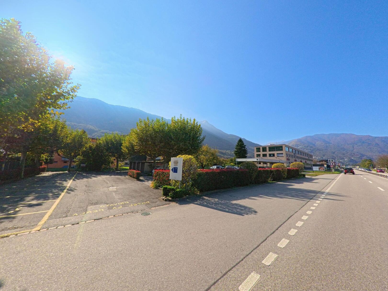 Entlang einer breiten Strasse in Sant’Antonino (6592, Kanton Tessin) erstrecken sich Parkflächen, Hecken und Gebäude vor der Kulisse bewaldeter Berghänge unter klarem Himmel.