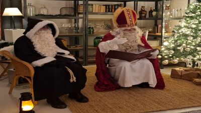In Muttenz (4132, Kanton Baselland) sitzen Samichlaus und Schmutzli in traditioneller Kleidung in einem festlich geschmueckten Raum und lesen aus einem Buch neben dem Weihnachtsbaum.