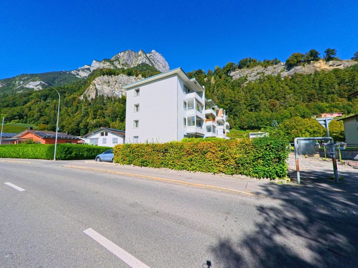Ein Mehrfamilienhaus in Sargans (7320, Kanton St. Gallen) steht am Strassenrand mit Blick auf bewaldete Felsen und sonnige Berghaenge.