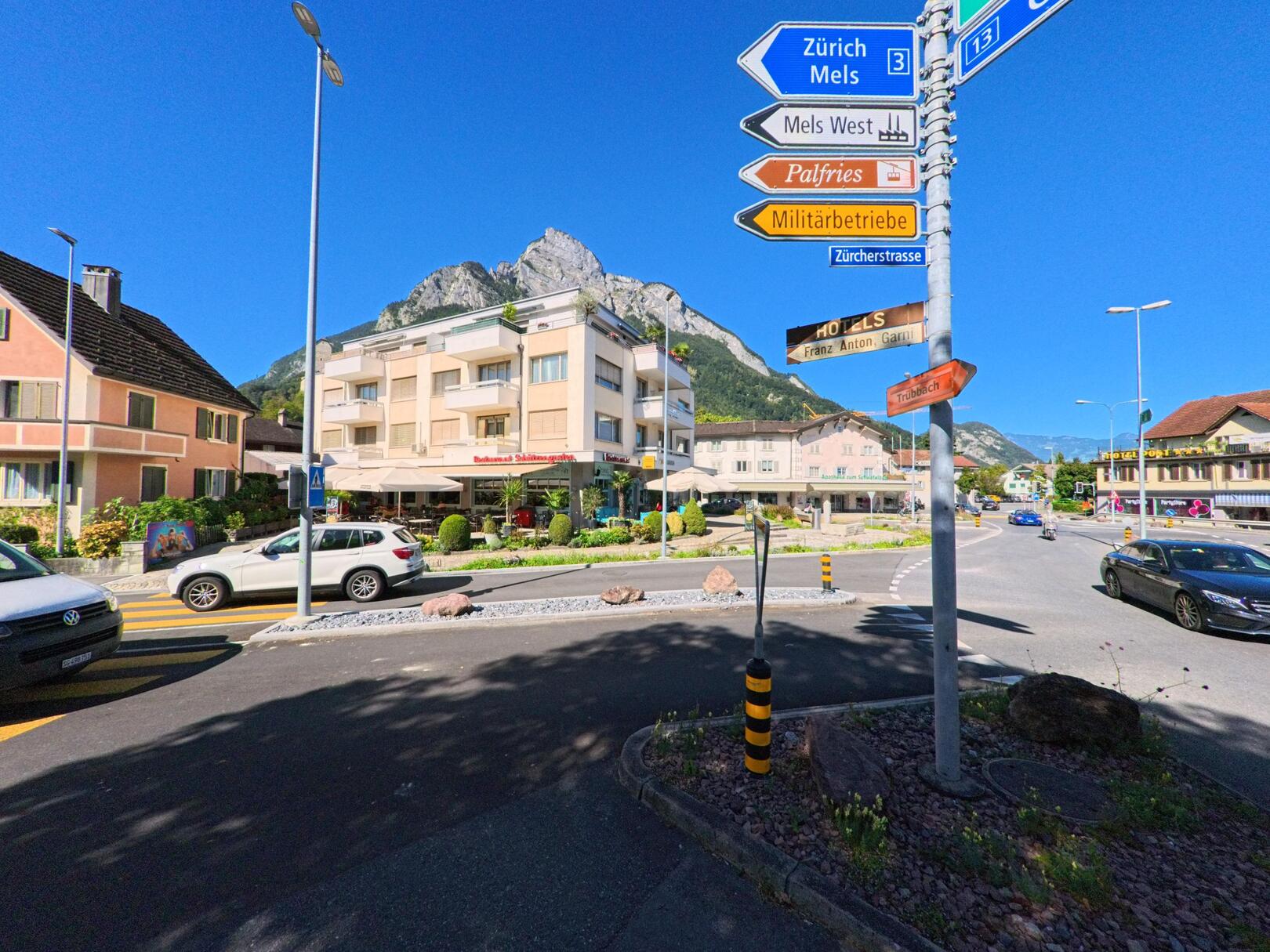 In Sargans (7320, Kanton St. Gallen) führt ein Fussgängerstreifen zu einem Gebäude mit Restaurantterrasse, während im Hintergrund markante Felsen aufragen.