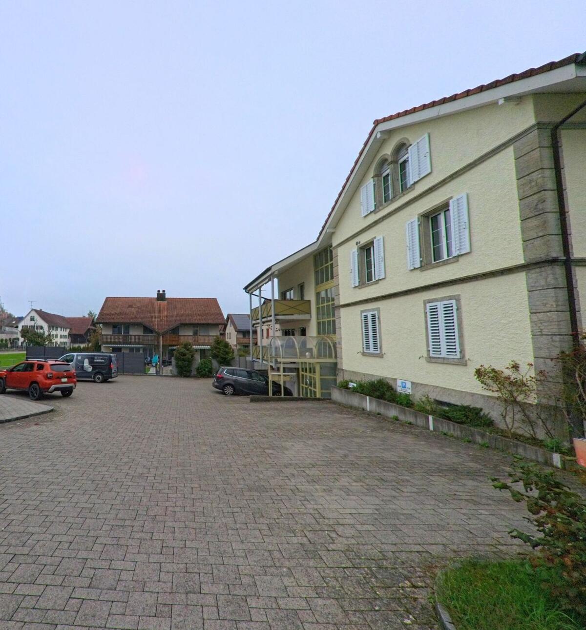 Ein helles Mehrfamilienhaus in Sarmenstorf (5614, Kanton Aargau) mit weissen Fensterläden und mehreren parkierten Autos steht an einem gepflasterten Hof zwischen weiteren Wohngebäuden.