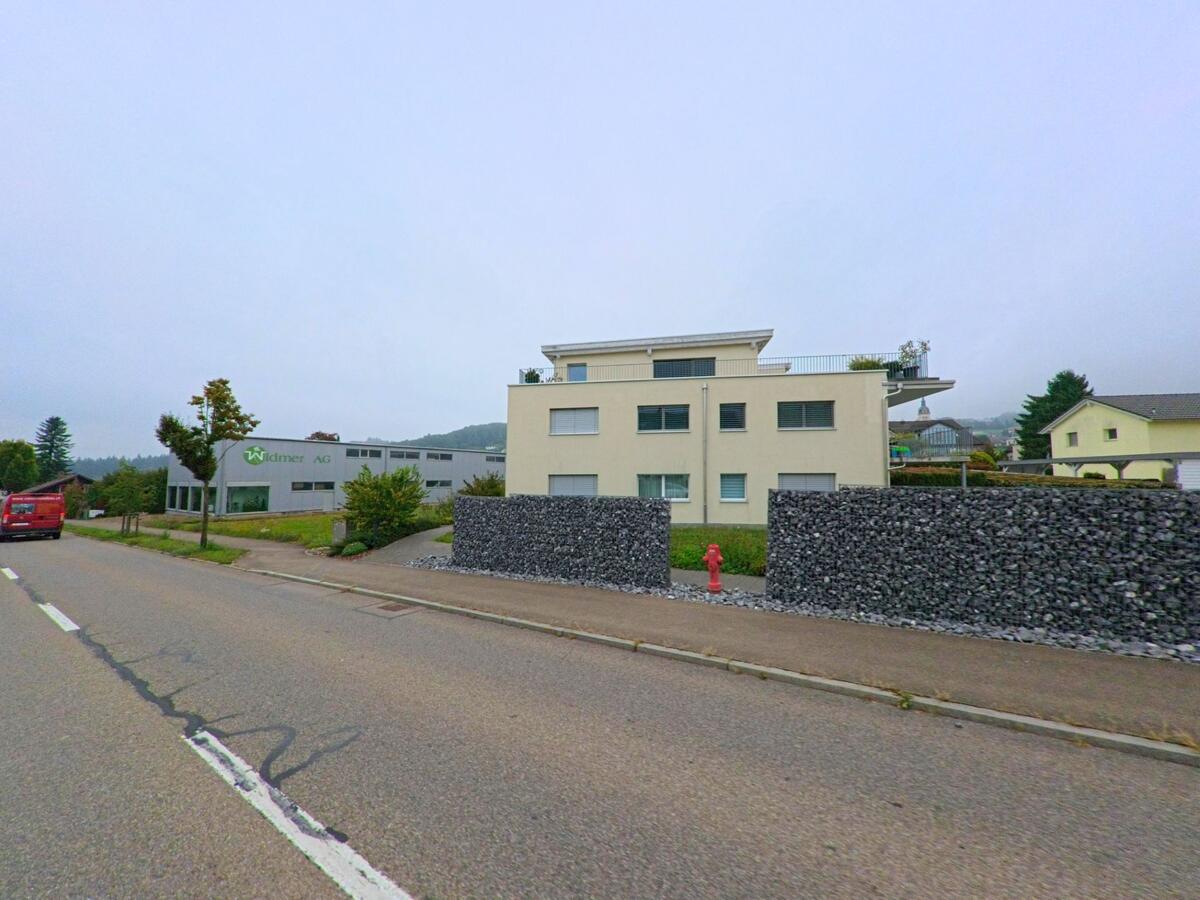Ein modernes Wohn- und Geschäftsgebäude in Sarmenstorf (5614, Kanton Aargau) steht an einer Hauptstrasse, umgeben von gepflegten Steinmauern und Grünflächen.