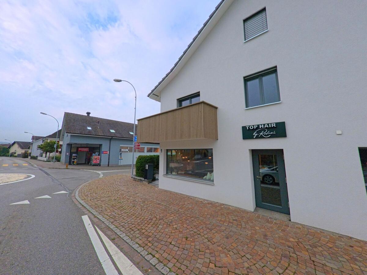 Ein modernes Wohnhaus in Sarmenstorf (5614, Kanton Aargau) mit einem Friseursalon im Erdgeschoss liegt an einer gepflasterten Strassenecke mit weiteren Gebäuden im Hintergrund.