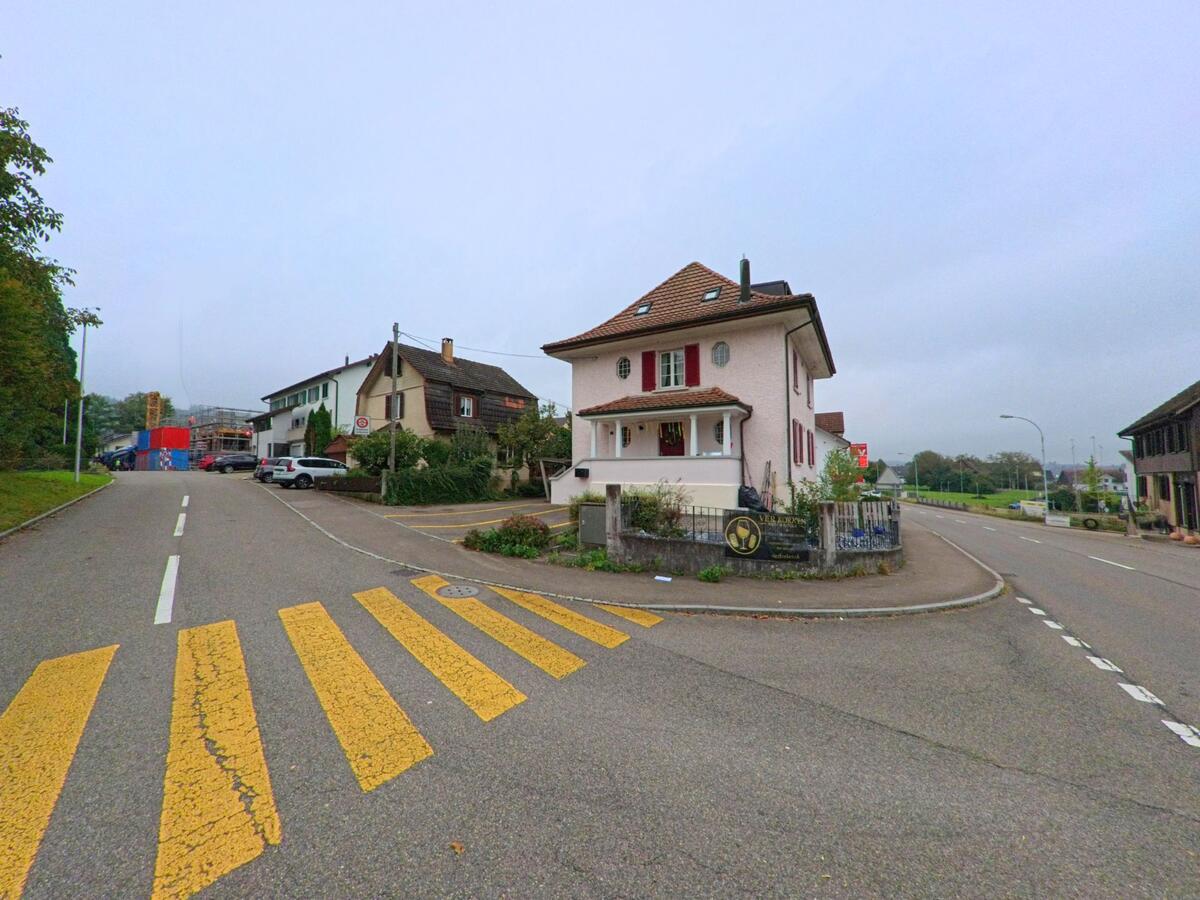 Ein Haus mit roten Fensterläden steht an einer Strassengabelung in Sarmenstorf (5614, Kanton Aargau), flankiert von weiteren Wohngebäuden und einem gelb markierten Fussgängerstreifen.