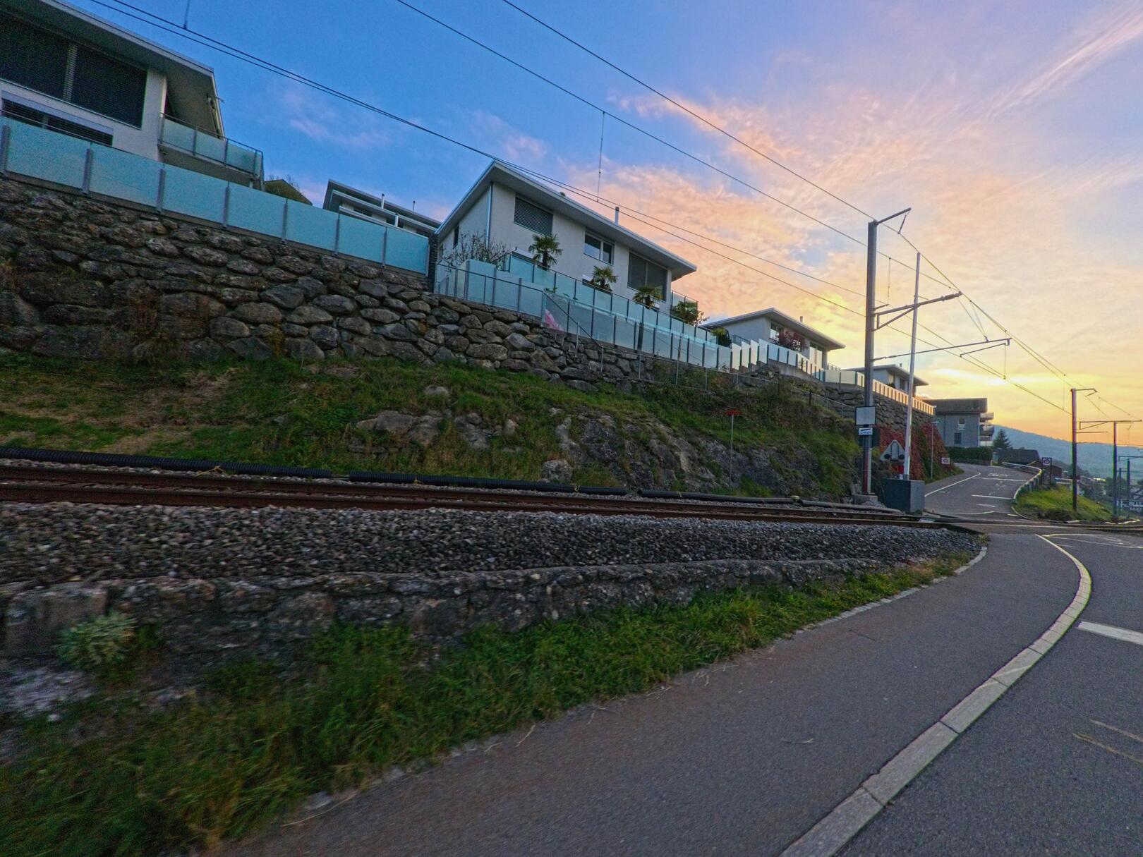 In Sattle (6417, Kanton Schwyz) verläuft ein Gehweg entlang der Gleise, während oberhalb moderne Wohnhaeuser auf einer gestuften Steinmauer im Abendlicht stehen.