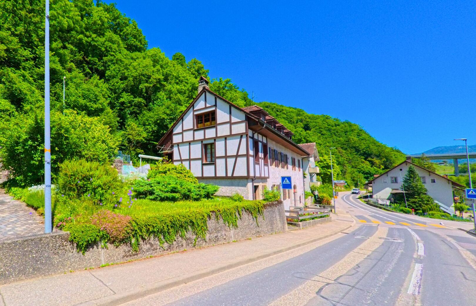 In Saugen (2535, Kanton Bern) ist ein Fachwerkhaus an einer kurvigen Strasse zu sehen, die von grünen Hügeln umgeben ist, mit einer Brücke im Hintergrund.