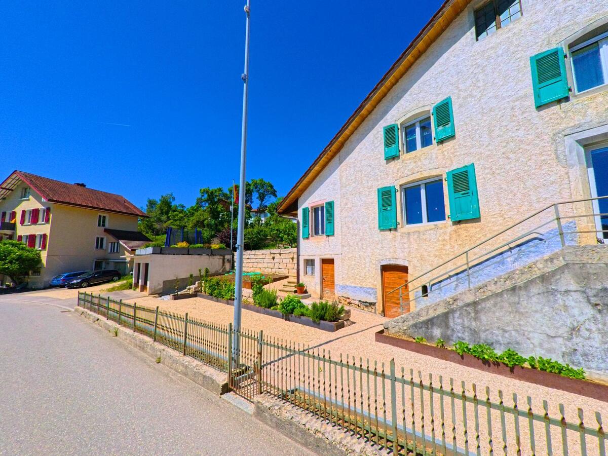  In Saugen (2536, Kanton Bern) steht ein Gebäude mit grünen Fensterläden, das von einer gepflegten Kiesfläche und einem schmiedeeisernen Zaun umgeben ist.