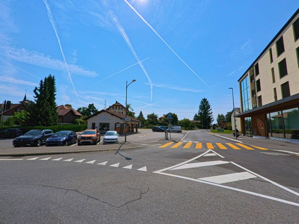 Ein Fussgaengerstreifen fuehrt in Savigny (1073, Kanton Waadt) von einem modernen Gebaeude zu einem kleinen Haus mit Geschaeft, daneben parken mehrere Autos.