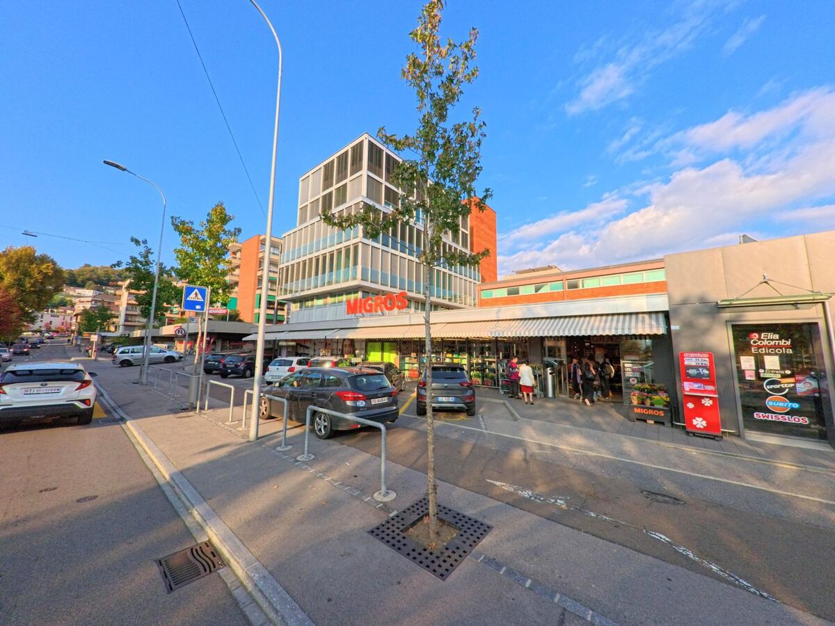 Vor einem Einkaufszentrum in Savosa (6942, Kanton Tessin) erstrecken sich Parkplätze, Gehwege und moderne Gebäude bei lebhaftem Betrieb im Tageslicht.