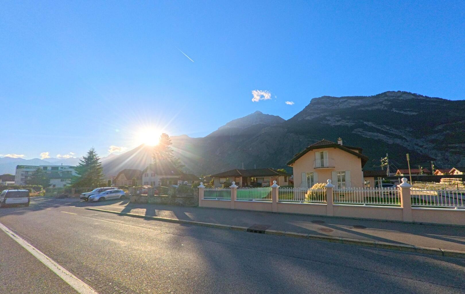 Ein Einfamilienhaus mit Gartenzaun steht in Saxon (1907, Kanton Wallis) an einer Strasse, waehrend die Sonne hinter den Bergen untergeht.