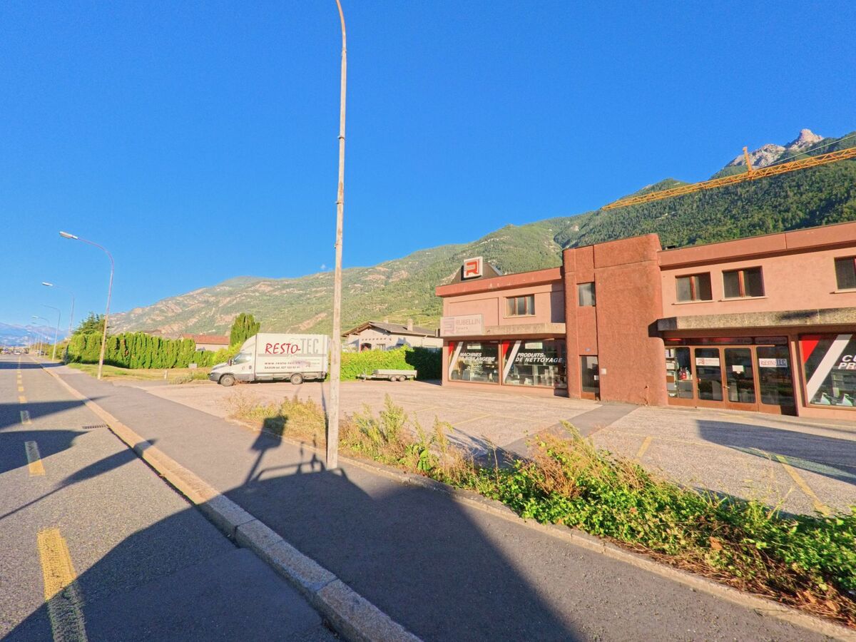 Ein Gewerbegebaeude mit grossen Schaufenstern steht in Saxon (1907, Kanton Wallis) an einer breiten Strasse mit Blick auf die Berge.