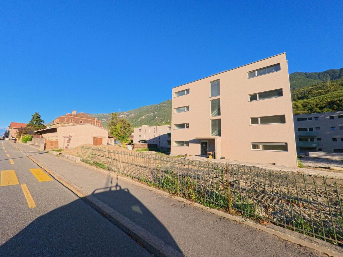 Ein modernes Wohnhaus steht in Saxon (1907, Kanton Wallis) neben einem aelteren Gebaeude an einer Strasse mit Gehweg.