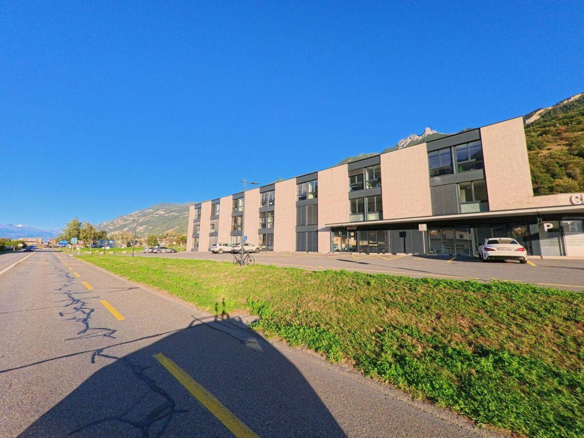 Ein modernes Gebaeude mit Geschaeften und Wohnungen steht in Saxon (1907, Kanton Wallis) direkt an einer Strasse mit Radweg.
