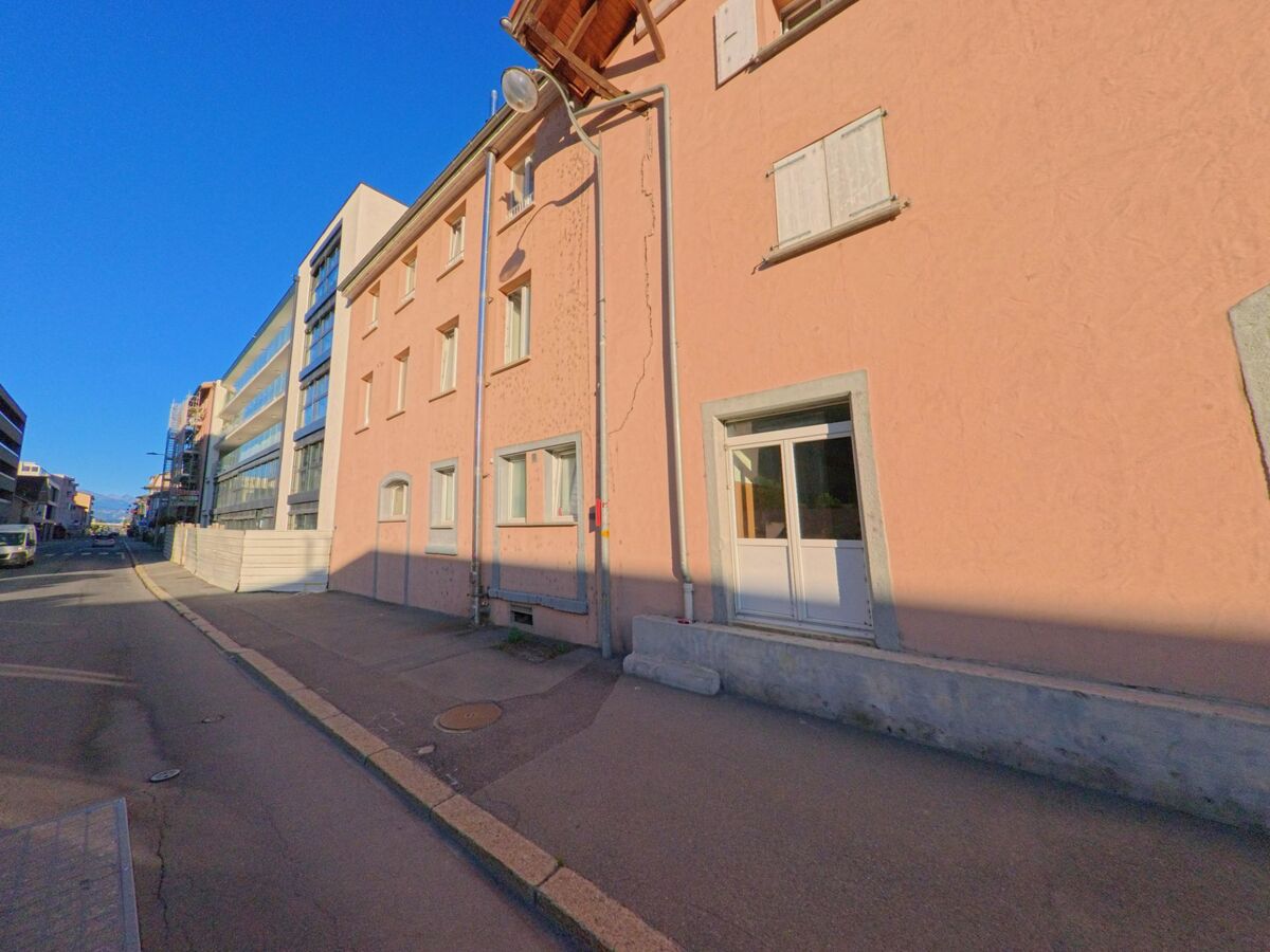 Eine enge Strasse in Saxon (1907, Kanton Wallis) verläuft zwischen einem rosa Wohnhaus und modernen Gebaeuden mit Glasfassaden.