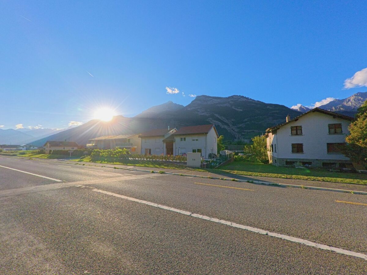 Am Ortsrand von Saxon (1907, Kanton Wallis) stehen Wohnhaeuser, waehrend die Sonne hinter den Bergen im Abendlicht versinkt.