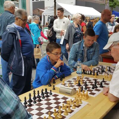 An einem Tisch in Buchs SG (9470, Kanton St.Gallen) spielen mehrere Personen Schach im Freien, waehrend Passanten das Geschehen aufmerksam verfolgen.