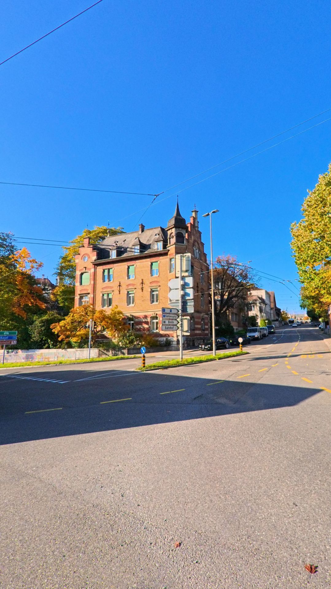 An einer breiten Kreuzung mit Blick auf ein historisches Backsteingebaeude und herbstlich gefaerbte Baeume zeigt sich ein Stadtabschnitt von Schaffhausen (8200, Kanton Schaffhausen) bei klarem Himmel.
