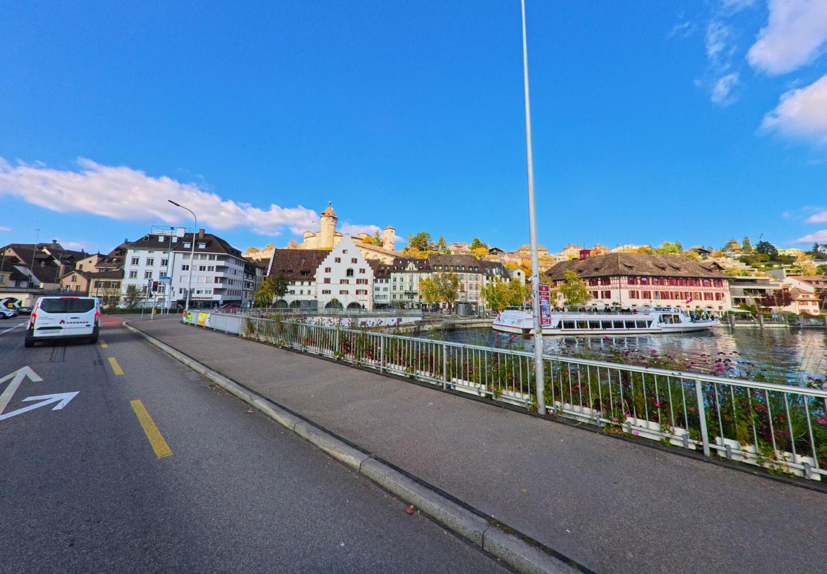 Entlang des Rheinufers mit Blick auf historische Fassaden und die Munot zeigt sich Schaffhausen (8200, Kanton Schaffhausen) bei klarem Himmel mit einem Schiff auf dem Fluss.